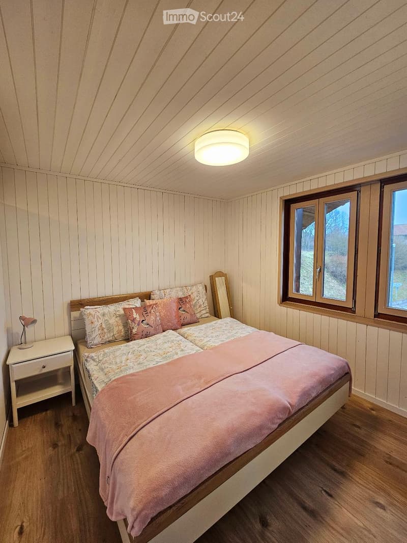 Cozy bedroom with wooden walls and ceiling, featuring a double bed with pink bedding, a nightstand with a lamp, and a mirror leaning against the wall. Large windows offer natural light.