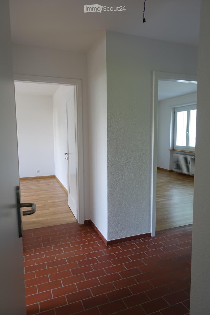Empty hallway with red tiled floor and wooden flooring in adjacent rooms, white walls, and doors leading to other spaces.