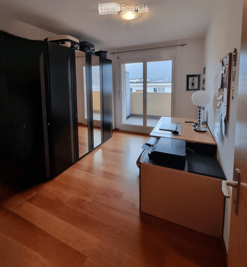 A furnished room with wooden flooring, a desk with a laptop, printer, and lamp, a large mirror, and a picture frame on the wall. A sliding glass door leads to a balcony.