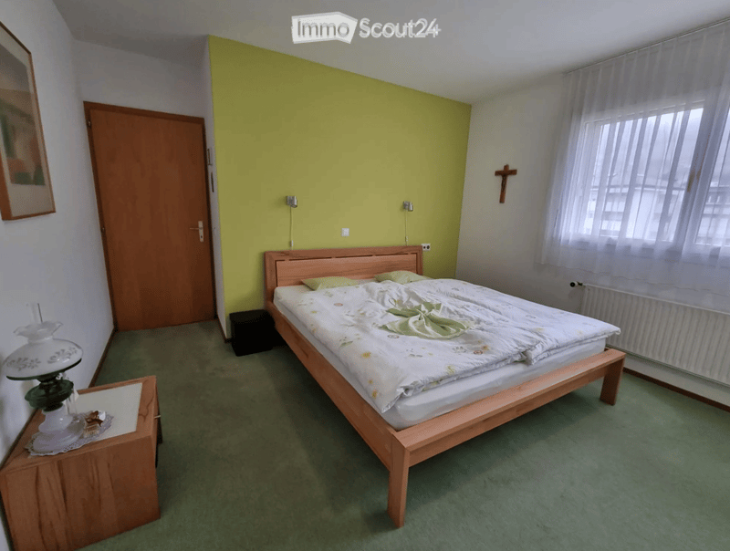 Bedroom with a wooden bed, nightstand, lamp, and a picture frame on the wall. Green carpeted floor and a window with white curtains.