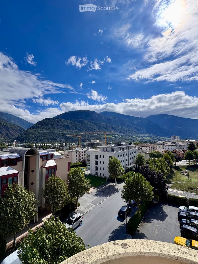 Balcony view overlooking a residential area with buildings, parked cars, trees, and distant mountains under a partly cloudy sky.