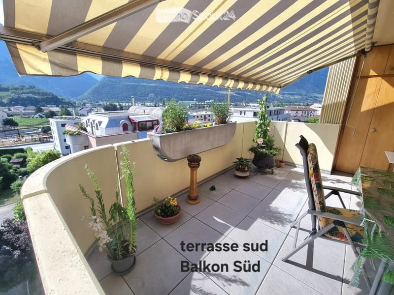 Balcony with tiled floor, potted plants, a chair, and a table, offering a scenic mountain view.