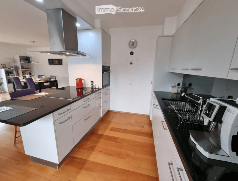 Modern kitchen with white cabinets, black countertops, and a central island. Includes a sink, dishwasher, oven, and a dining area with chairs and a table.