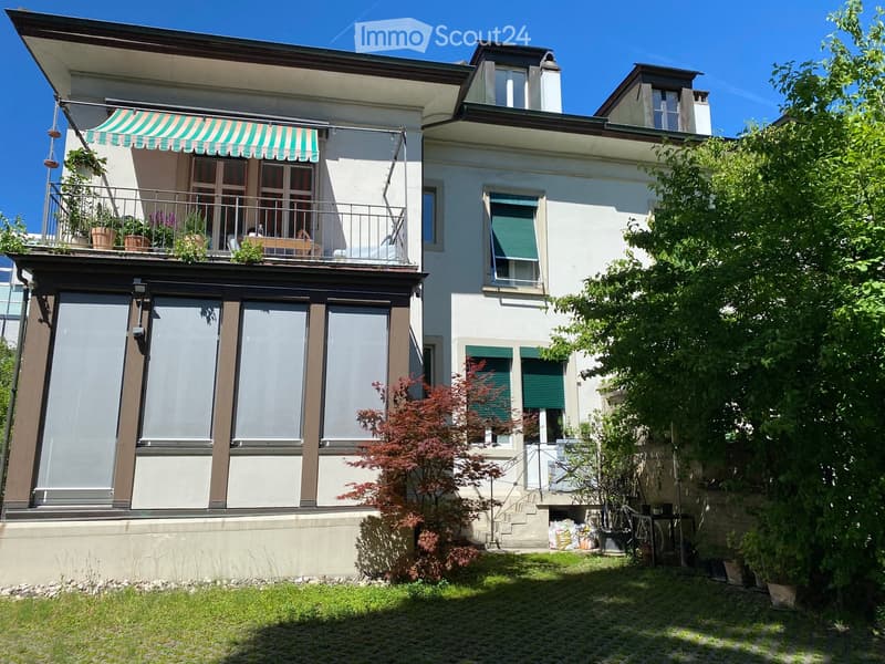Two-story house with a balcony, green shutters, and a small garden area with plants and a tree.