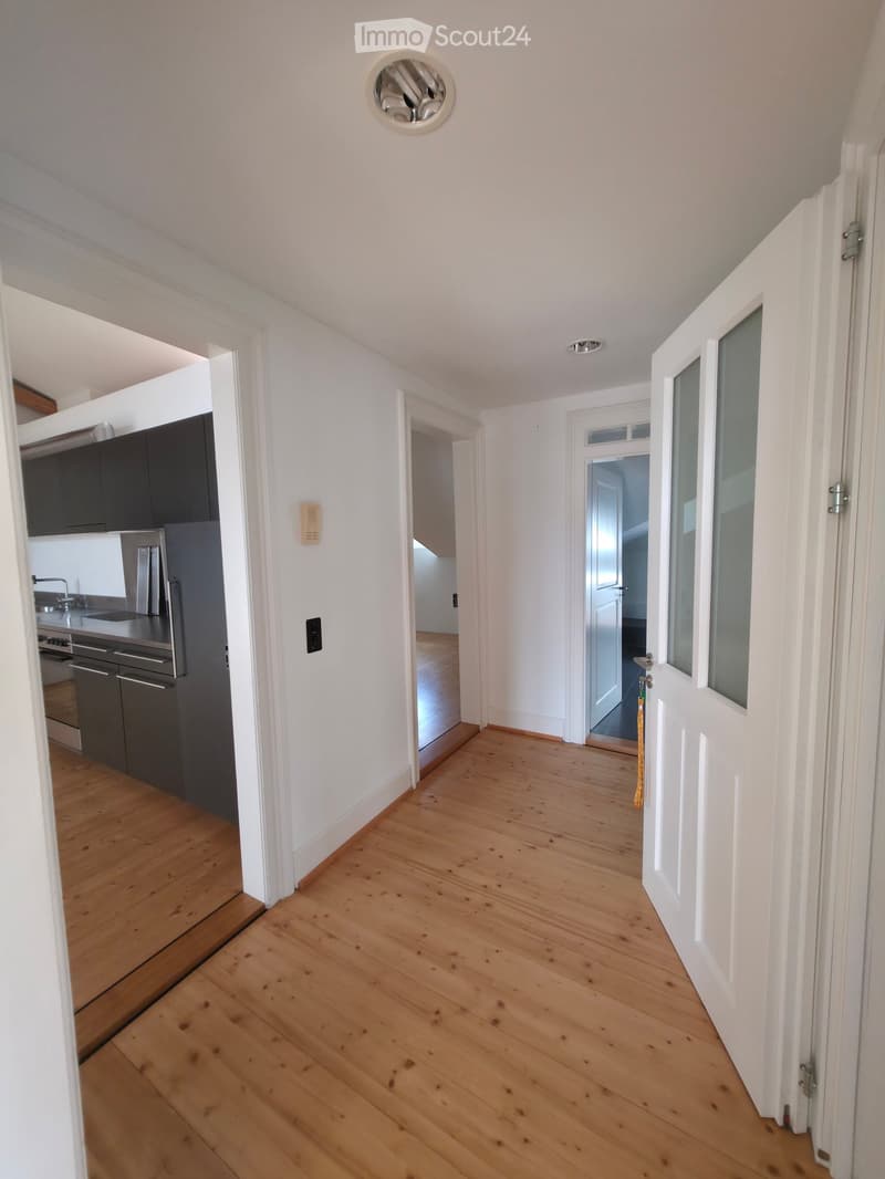 Bright hallway with wooden flooring, white walls, and multiple doors leading to different rooms, including a kitchen area visible on the left.