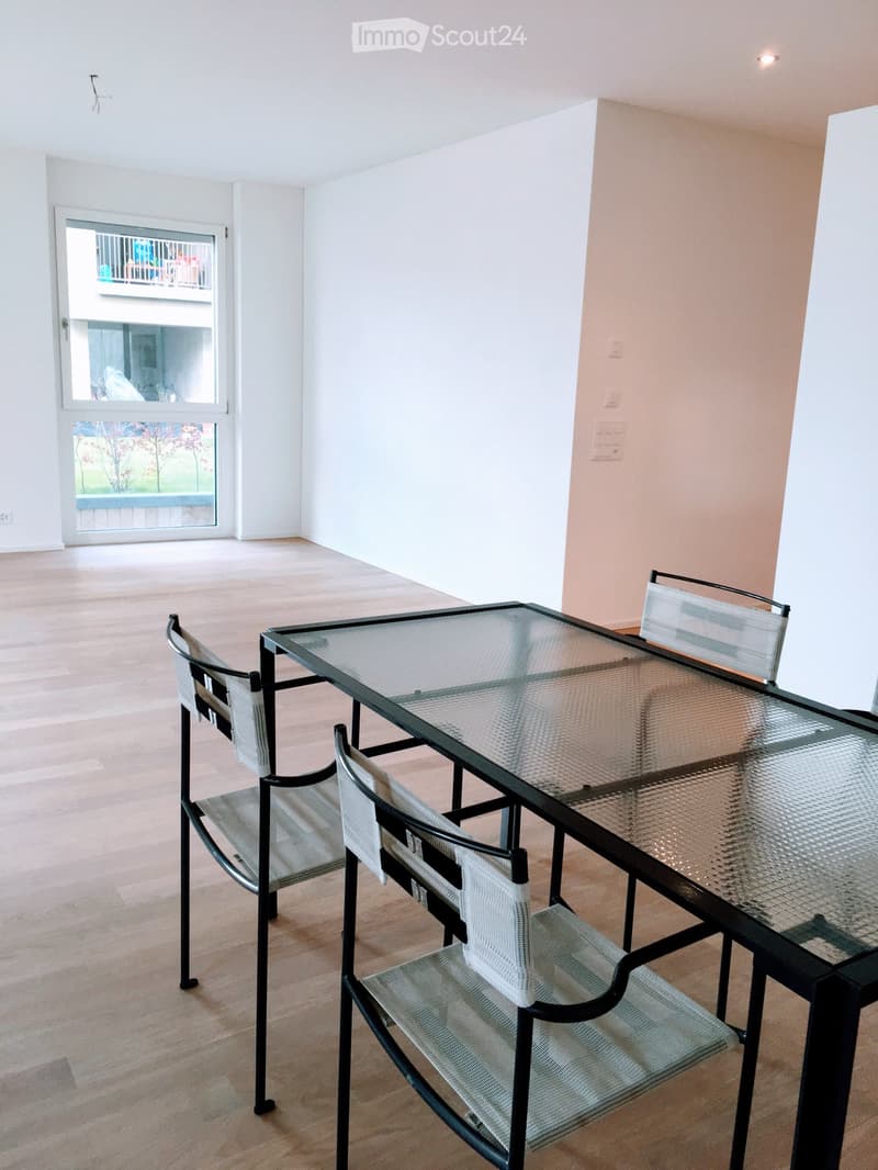 Bright, empty room with wooden flooring, a glass-top dining table with four white chairs, and a large window leading to a balcony.