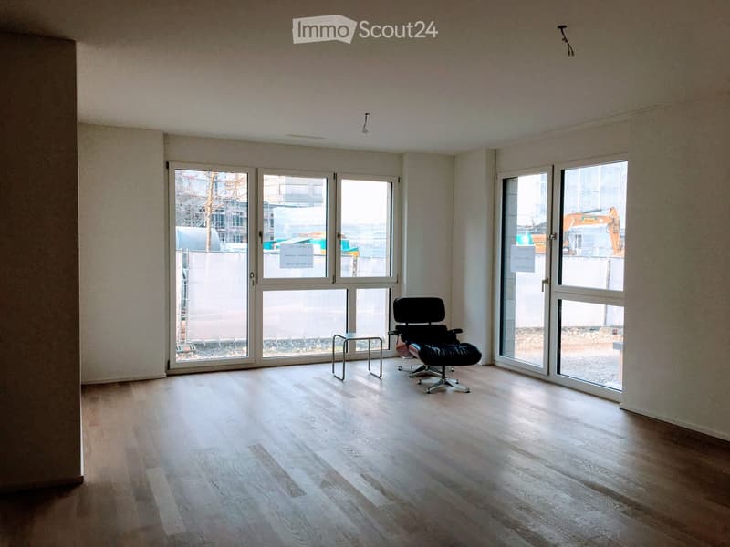 Bright, unfurnished room with wooden flooring, large windows, and a single black chair and stool.