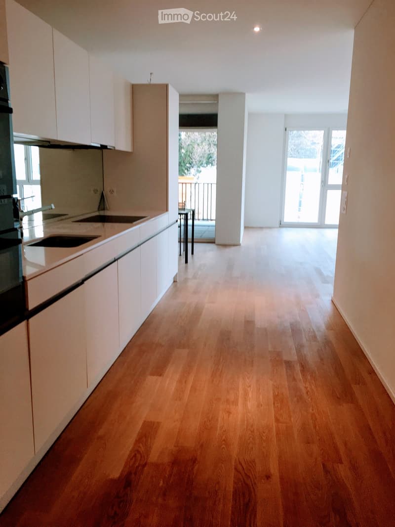 Modern kitchen with white cabinets, a double sink, and a sleek oven. The floor is made of light wooden planks, and the space opens to a balcony area with a small table and chairs.