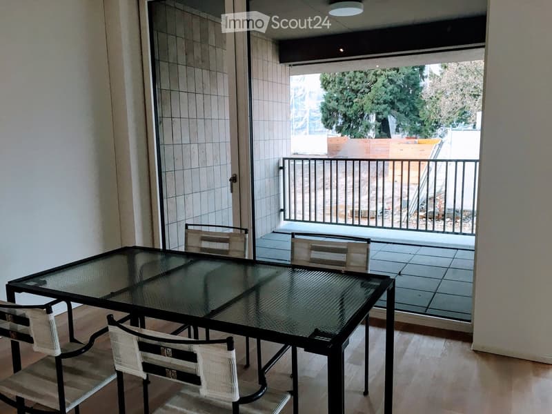 Dining area with a glass table and four white chairs, leading to a tiled balcony with a metal railing and outdoor view.