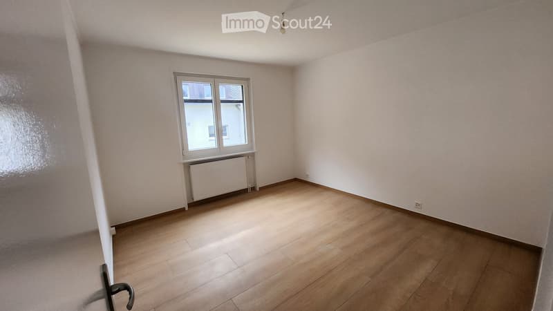 Empty room with wooden flooring, white walls, a window with white frame, radiator, and a light fixture on the ceiling.