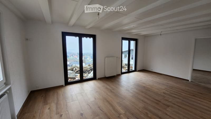 Empty room with wooden flooring, white walls, and large windows offering a view of the outside area.
