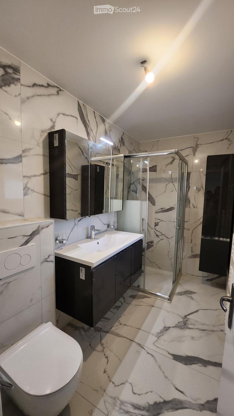 Modern bathroom with marble tiles, a white toilet, a double sink with a modern faucet, a large mirror, and a glass-enclosed shower area.