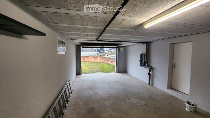 Empty concrete-floored corridor with white walls, a small window, a door, and visible pipes. No furniture or equipment present.