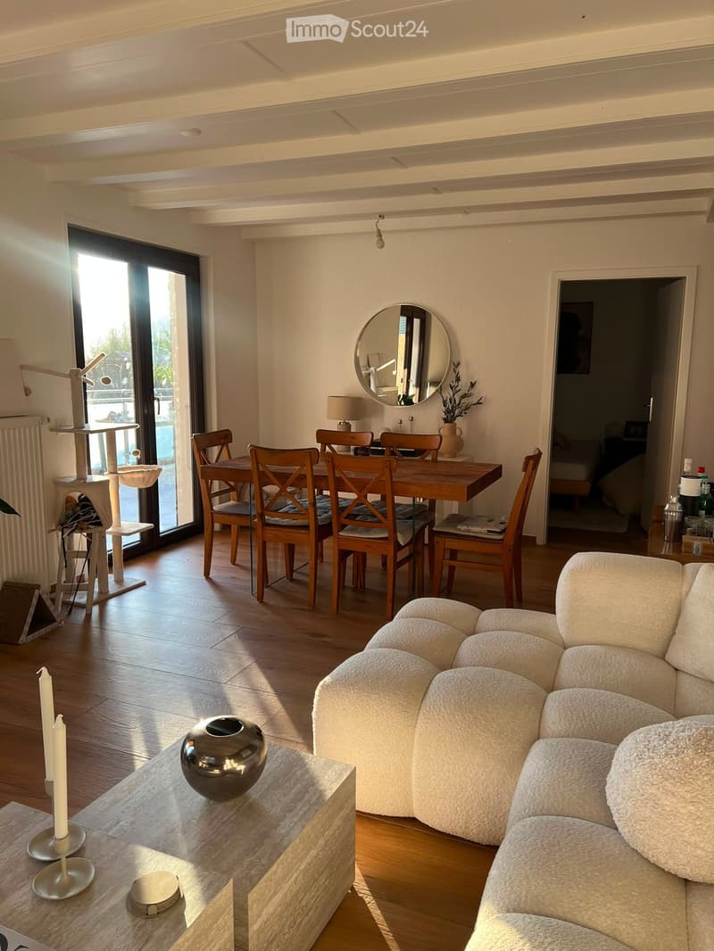 A modern, well-lit living and dining area with a white sectional sofa, a coffee table with candles and a decorative sphere, a cat tree, a dining table with six wooden chairs, a round mirror, and a small table lamp.
