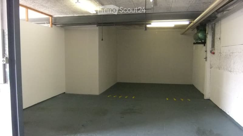 Empty garage with concrete floor, white walls, and fluorescent lighting. A small window on the left and a green air conditioning unit on the right wall.