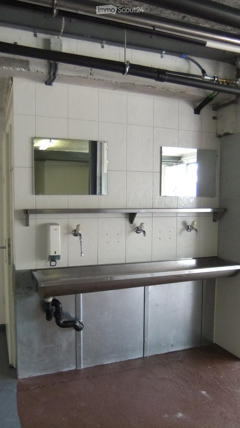 Industrial-style bathroom with stainless steel sink, multiple faucets, mirrors, and a soap dispenser mounted on tiled walls.