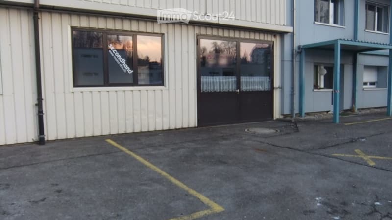 Exterior view of a multi-story building with white siding, large windows, and a covered entrance area. The ground floor has a large door and a window with a sign reading "secondhand."
