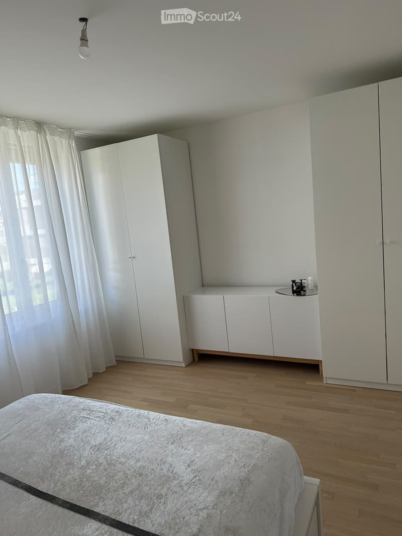 Modern bedroom with a large bed, white wardrobes, a small table with decorative items, and sheer curtains on the window.