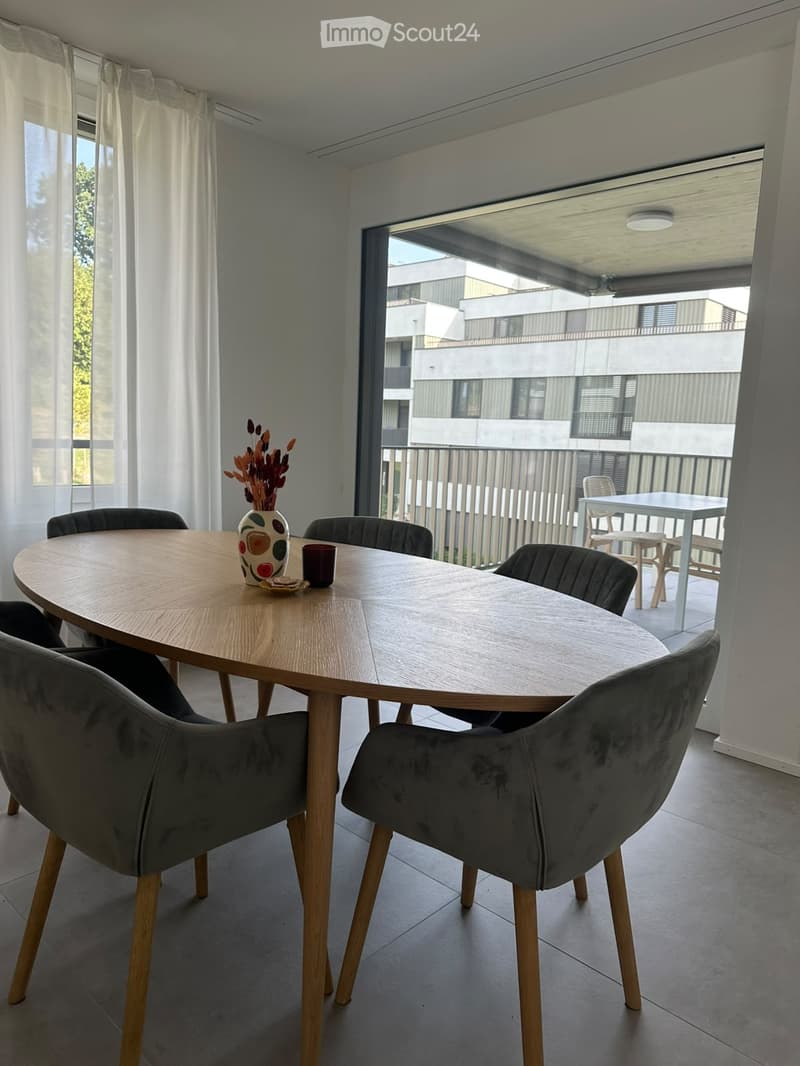Dining area with a round wooden table, four grey upholstered chairs, a decorative vase with dried flowers, and a cup on the table. Large windows and sliding glass doors lead to a balcony with outdoor furniture.