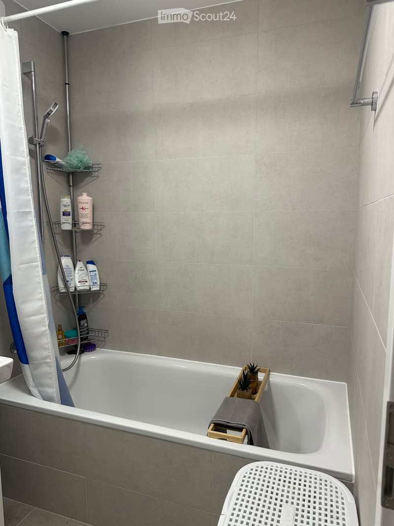 Modern bathroom with a bathtub, shower curtain, and a shelf holding various bottles and toiletries. A small wooden tray with plants and a folded towel sits in the bathtub.