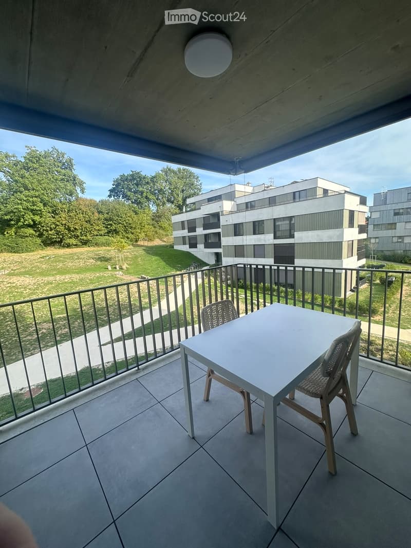 Balcony with a white table and two chairs, overlooking a green area and modern buildings.