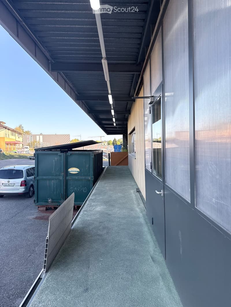 A covered outdoor corridor with a concrete floor, metal roof supported by white lights, and grey walls with windows. Green dumpsters are parked nearby, and a silver car is visible in the background.