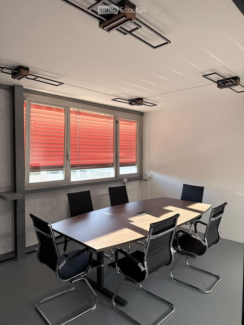 Modern conference room with a large rectangular table surrounded by black chairs, large windows with red blinds, and recessed lighting on the ceiling.