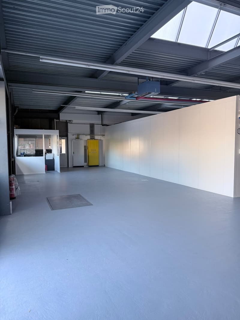 Empty room with white walls, grey floor, and industrial-style ceiling with exposed beams and lighting. Features a small reception area with a desk and chairs, a yellow storage unit, and a fire extinguisher.