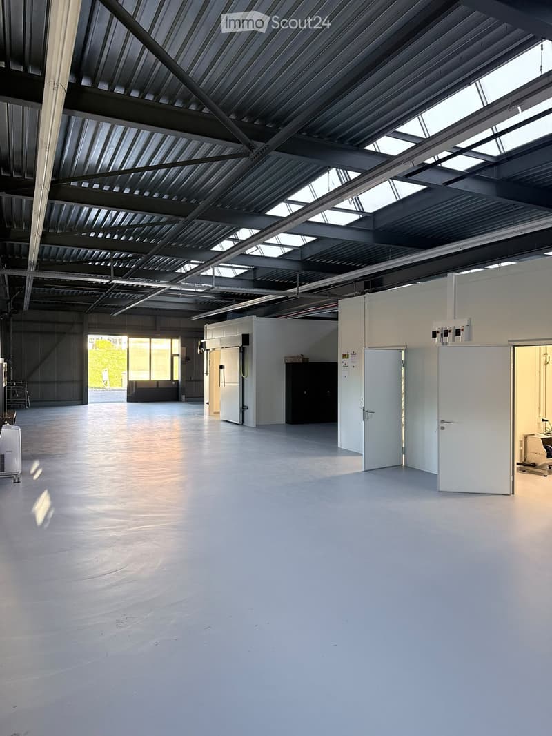 Large, empty industrial-style room with a high, metal-framed ceiling and skylights, white walls, and a smooth concrete floor. Minimal furnishings include a small cabinet and a few storage units.