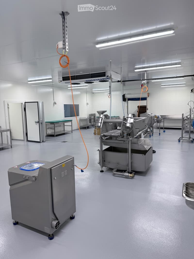 Clean, industrial kitchen with stainless steel equipment, tables, and machinery, featuring a large open space with white walls and a grey floor.