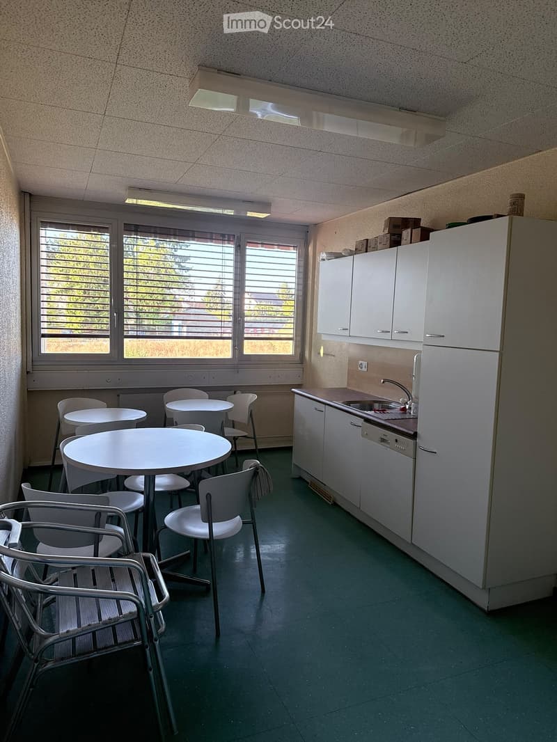 A small kitchen area with white cabinets, a refrigerator, sink, and dishwasher. There are several round tables and chairs arranged in the space, with a window offering a view of greenery outside.