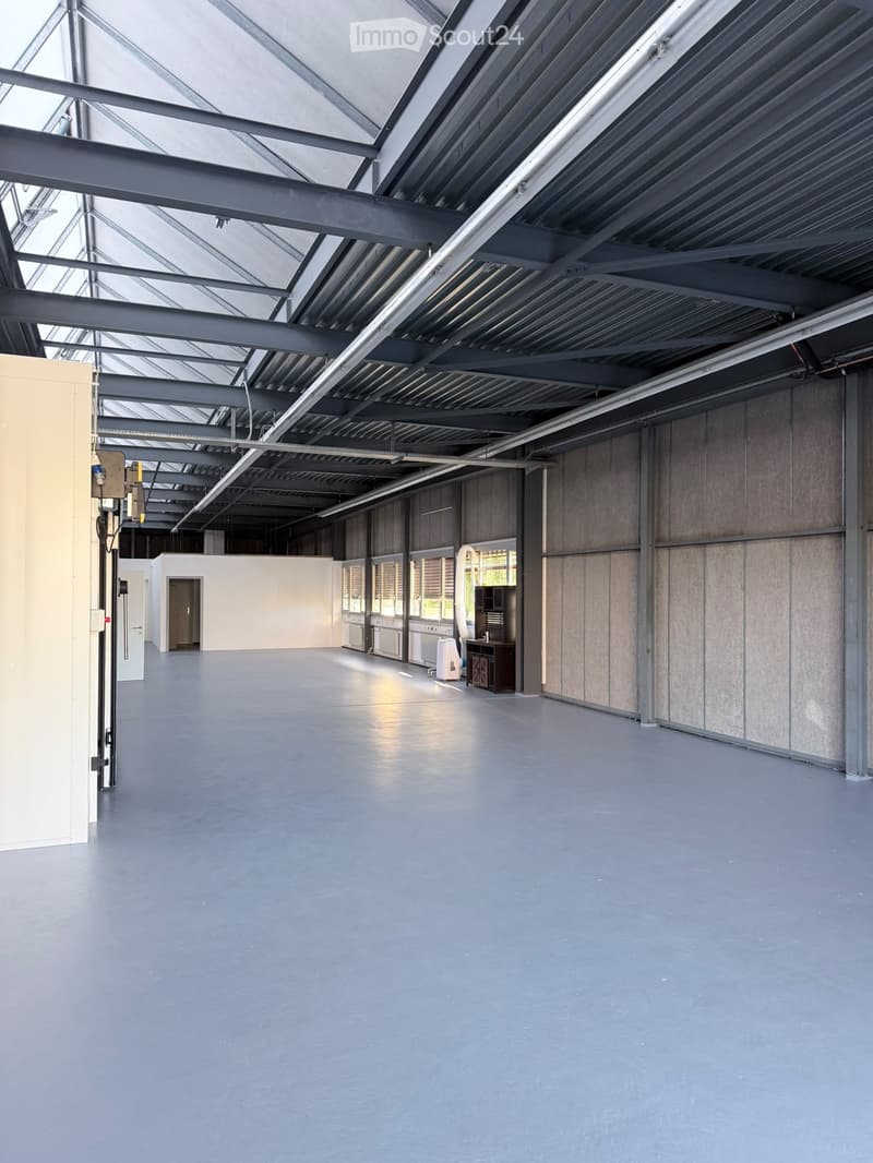 Large, empty industrial-style room with a high, metal-framed ceiling and large windows. The space features a smooth concrete floor and exposed metal beams, with minimal furnishings visible.