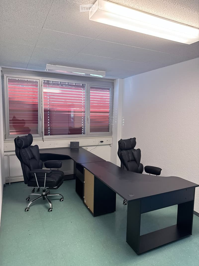 Office space with two black office chairs, a long black desk, and a small speaker on the desk. The room has large windows with blinds and fluorescent lighting.