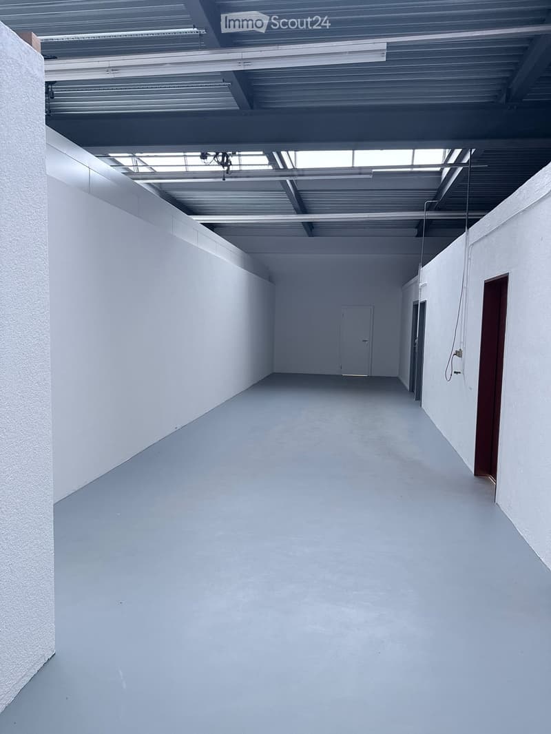 Empty, white-walled corridor with a single door at the far end and exposed ceiling beams and lighting fixtures.