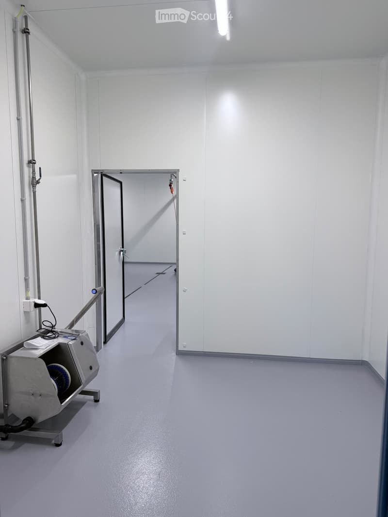Empty room with white walls, grey floor, and a metal door leading to another room. A small machine with a blue component is visible on the left side.