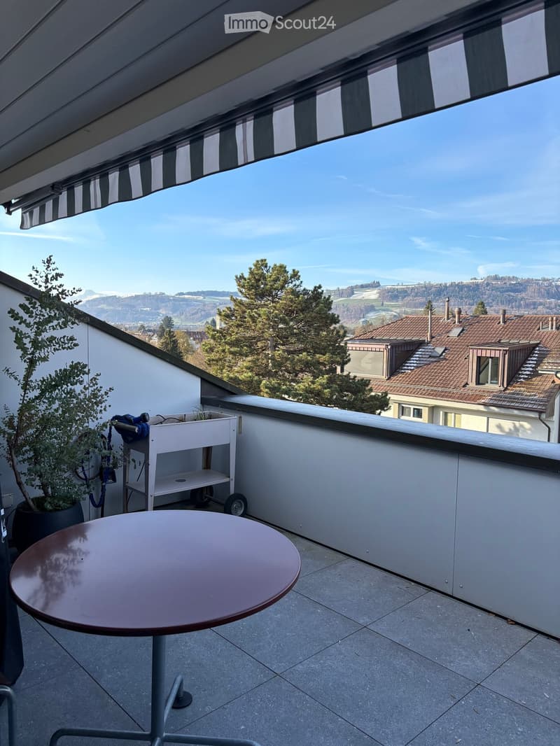 Balcony with a round table, a small cart, and a potted plant, offering a scenic view of the surrounding landscape and buildings.
