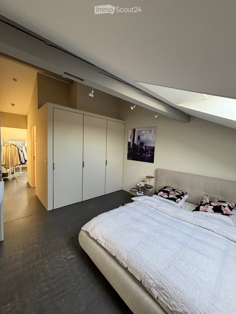 Modern bedroom with a large bed, nightstand, lamp, and a large wardrobe. The room features a slanted ceiling with skylight and dark flooring.