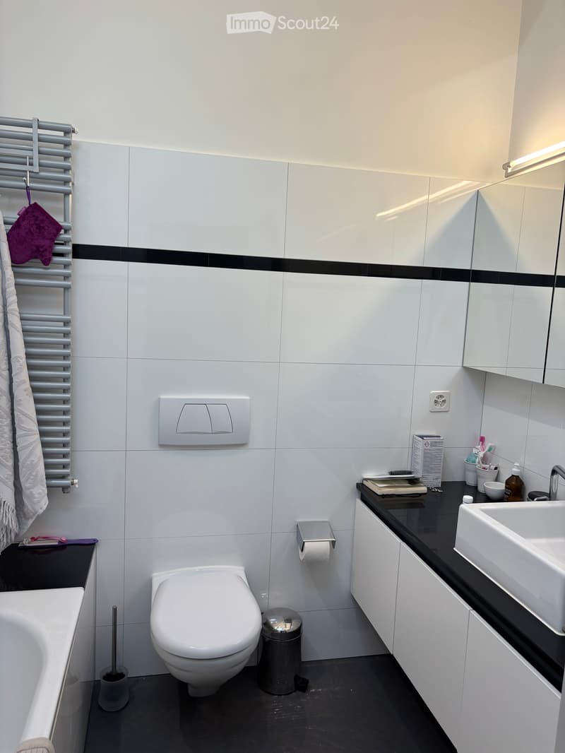 Modern bathroom with white tiles, black accents, a toilet, bathtub, sink, mirror, and cabinet. Includes a towel rack with towels, a toilet paper holder, and a trash can.