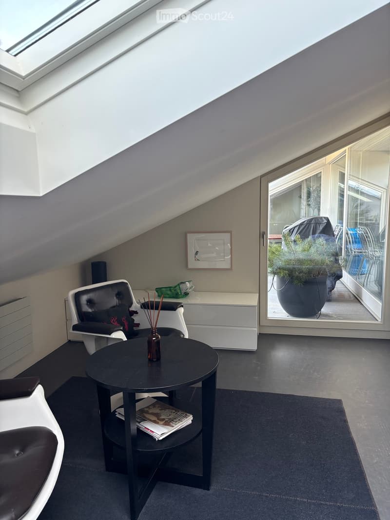 Modern, slanted-ceiling room with a black round table, white leather chair, and a large window leading to a balcony with a potted plant.