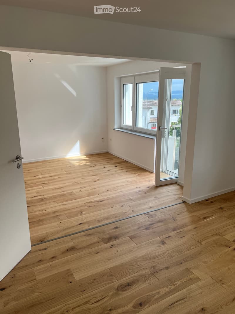 Empty room with light wooden flooring, white walls, and large windows with a sliding glass door leading outside.