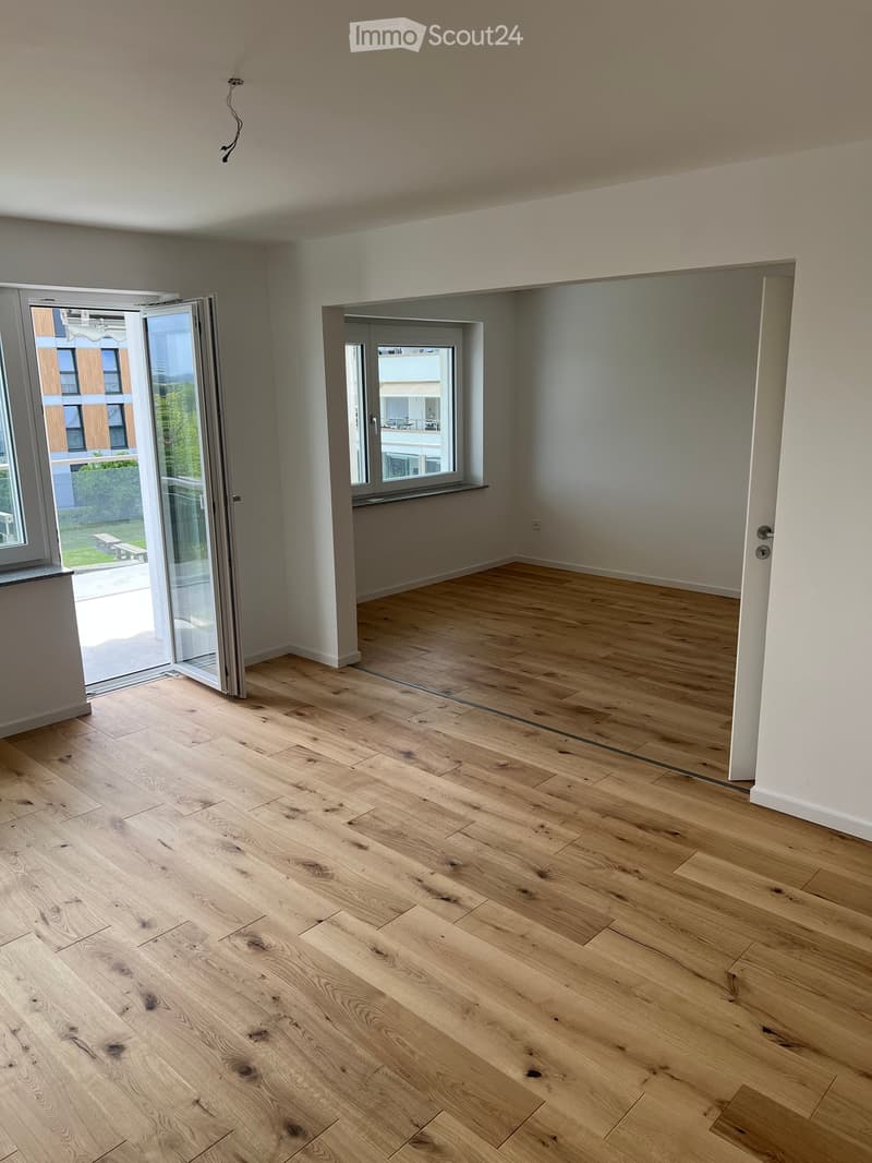Empty room with light wooden flooring, white walls, and large windows with sliding glass doors leading to a balcony.