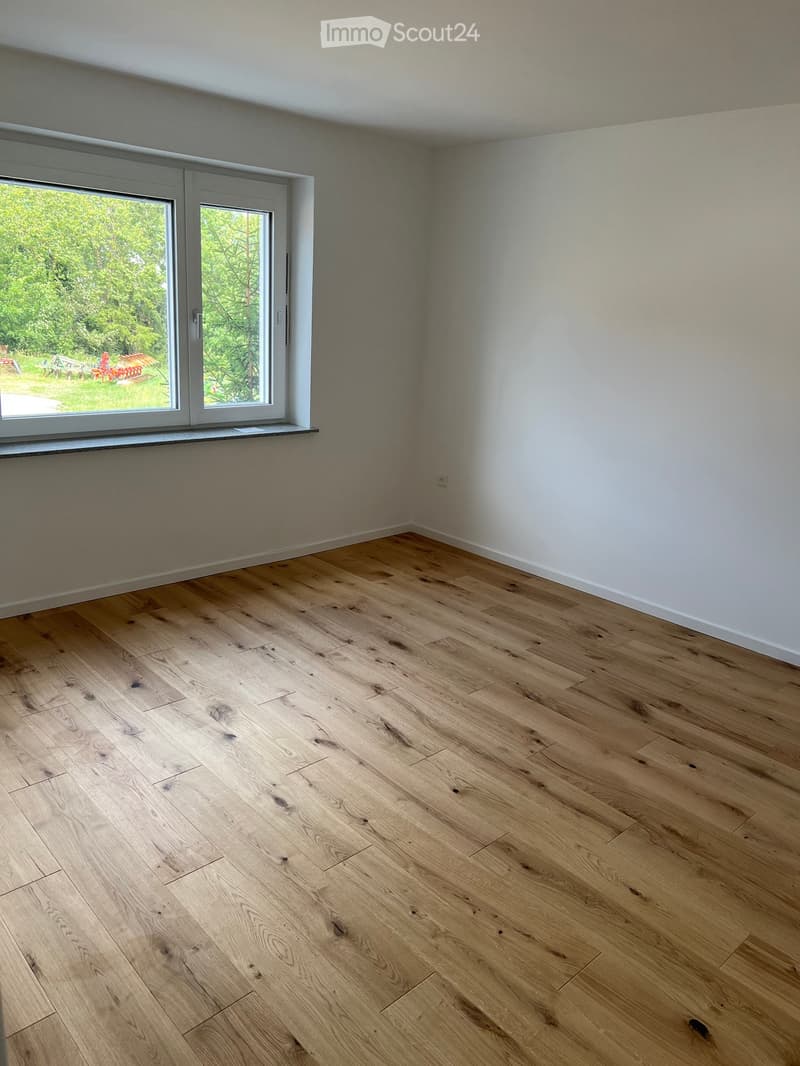 Empty room with light wooden flooring and white walls, featuring a large window with a view of greenery outside.