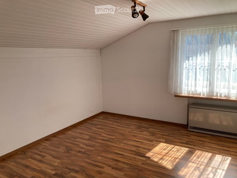 Empty room with wooden flooring, white walls, and a window with sheer curtains. A radiator is visible beneath the window, and a small shelf sits above it. The ceiling features recessed lighting fixtures.
