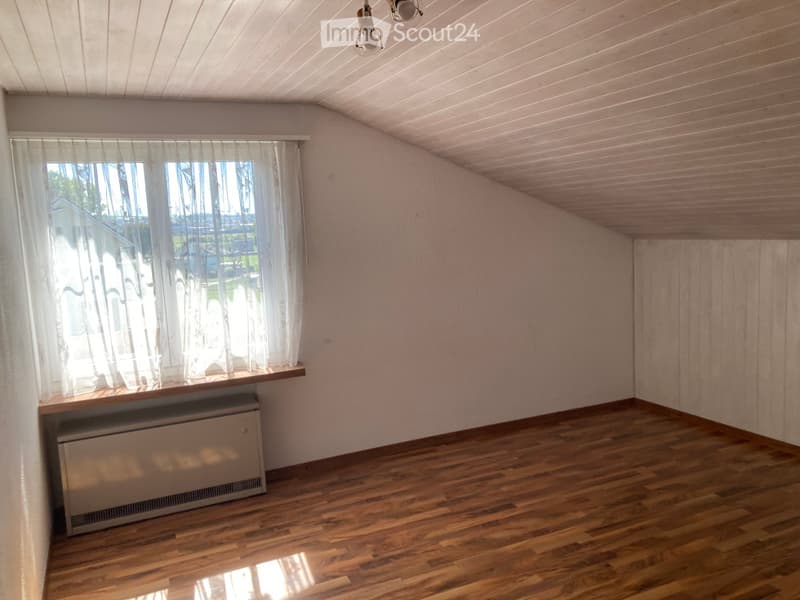 Empty room with wooden flooring, white walls, and a window with sheer curtains. A radiator is placed beneath the window, and the ceiling has a slanted design with recessed lighting.