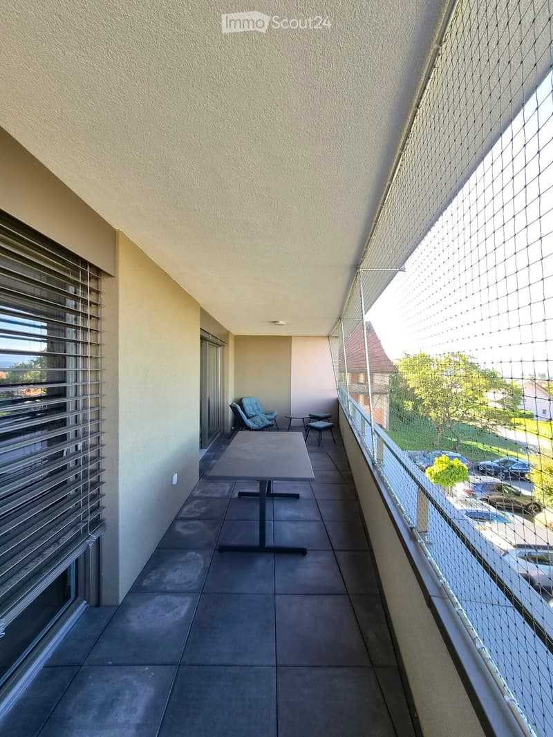 Balcony with tiled floor, table, chairs, and a baby chair, featuring a view of the street and parked cars.