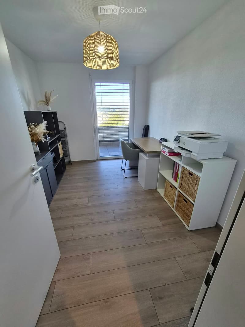 Bright, modern room with wooden flooring, white walls, and a hanging woven light fixture. Features a desk with a chair, a printer, shelves with books and wicker baskets, and a small black cabinet with decorative items.