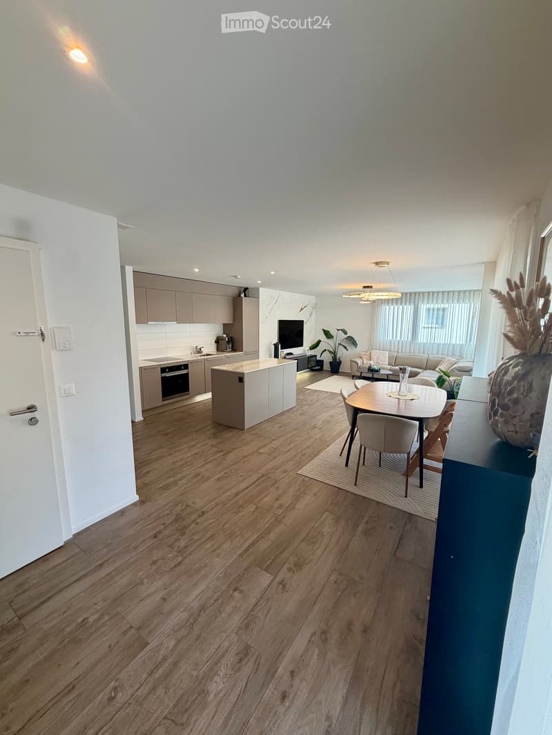 Open-plan living and dining area with wooden flooring, modern kitchen, sofa, dining table, and decorative vase.
