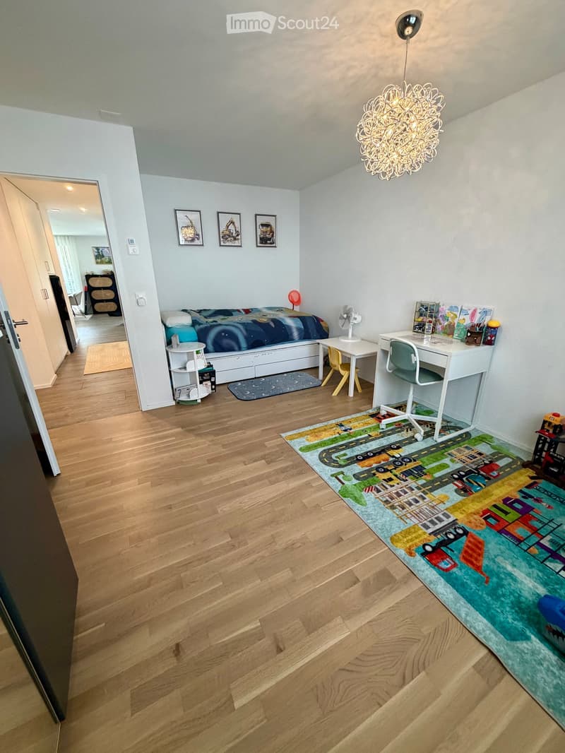 Bright bedroom with wooden flooring, a bed with colorful bedding, a desk with a chair, a small table, a fan, and a colorful play mat on the floor.