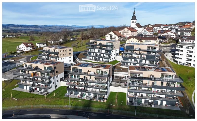 Aerial view of a modern residential complex with multiple apartment buildings, balconies, and solar panels on rooftops, surrounded by green fields and a town with a church tower in the background.