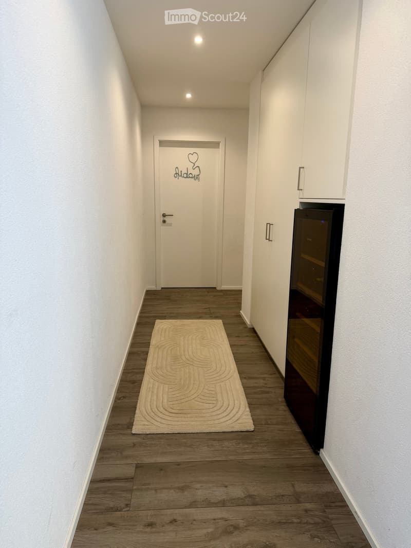 A narrow hallway with wooden flooring, white walls, a cream-colored circular rug, and a closed door with a heart design on it. A built-in cabinet with a black appliance is visible on the right side.